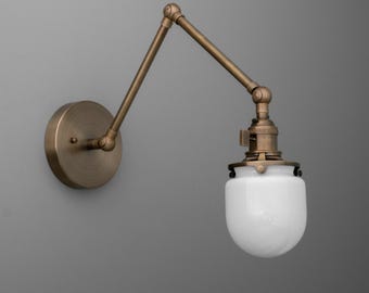 May include: A wall-mounted, adjustable brass-toned lamp with a white, teardrop-shaped glass shade. The lamp features a round base and an articulated arm for directional lighting. The design is reminiscent of vintage industrial lighting.