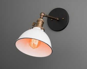 May include: A white, dome-shaped wall sconce with a black rim and a vintage-style lightbulb. The fixture has a bronze-colored arm and a black circular base. The light is mounted on a gray wall, showcasing an industrial design.