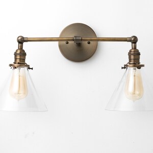 May include: A vintage-style wall sconce with two lights. The fixture has a round, bronze-colored backplate, a horizontal arm, and two downward-facing lights with clear, conical shades. Each light has a visible Edison-style bulb.