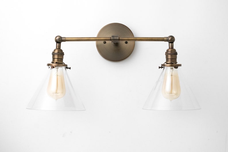 May include: A vintage-style wall sconce with two lights. The fixture has a round, bronze-colored backplate, a horizontal arm, and two downward-facing lights with clear, conical shades. Each light has a visible Edison-style bulb.