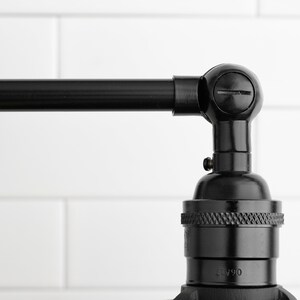 May include: Close-up of a black, industrial-style wall sconce. The sconce features a pivoting arm and a bulb socket. The finish is a dark, matte black, and the design is minimalist. The background is a white, tiled wall.