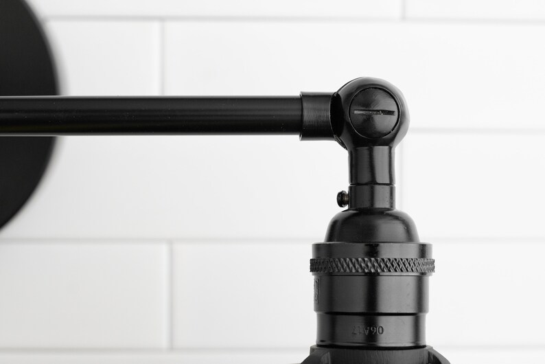 May include: Close-up of a black, industrial-style wall sconce. The sconce features a pivoting arm and a bulb socket. The finish is a dark, matte black, and the design is minimalist. The background is a white, tiled wall.