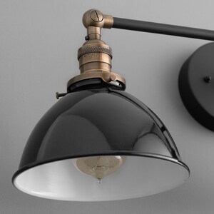 Puede incluir: Aplique de pared de estilo industrial negro y lat&oacute;n con dos pantallas de metal negro. Las pantallas tienen un interior blanco y est&aacute;n inclinadas hacia abajo. La aplique tiene un brazo de metal negro y una base redonda de metal negro.