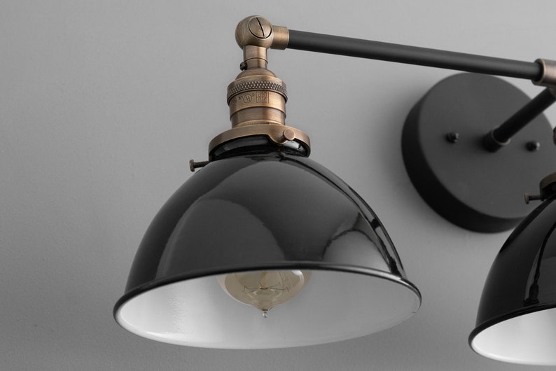 Puede incluir: Aplique de pared de estilo industrial negro y lat&oacute;n con dos pantallas de metal negro. Las pantallas tienen un interior blanco y est&aacute;n inclinadas hacia abajo. La aplique tiene un brazo de metal negro y una base redonda de metal negro.