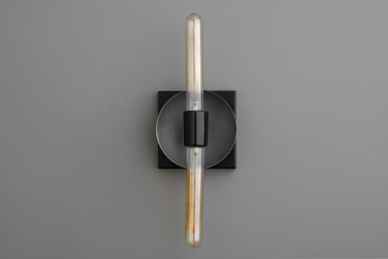 May include: A wall sconce featuring a matte black square base with a circular center plate. A central black cylindrical socket holds a long, clear tubular Edison-style light bulb with visible internal filaments, oriented vertically in the center.
