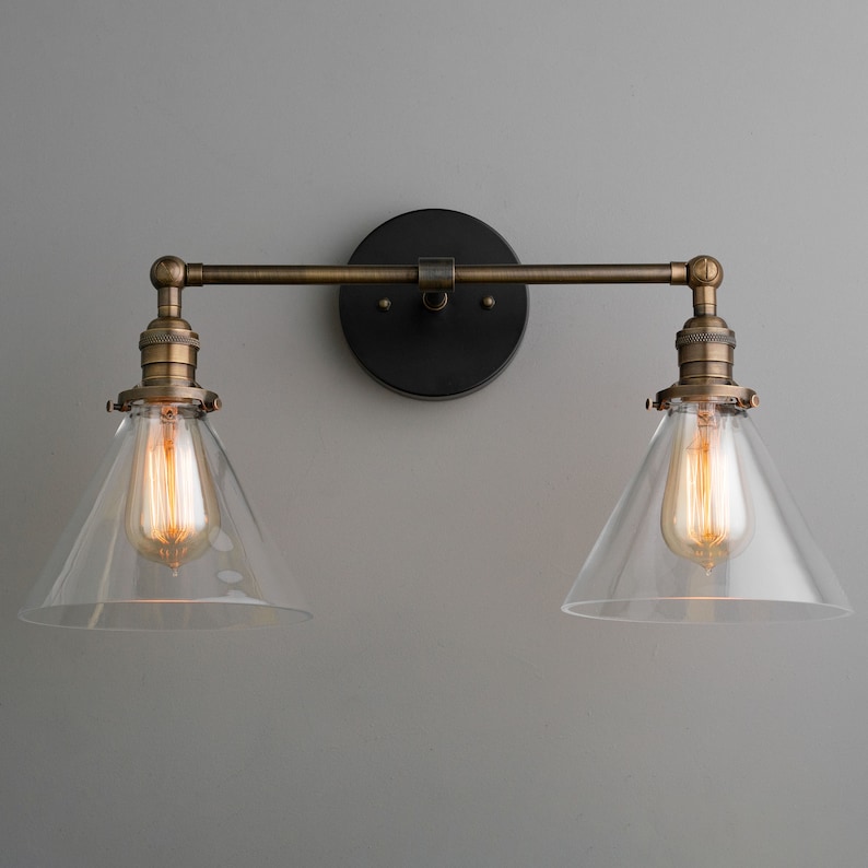 May include: A two-light wall sconce with a vintage industrial design. It features clear, cone-shaped glass shades and exposed Edison-style bulbs. The fixture has a dark bronze backplate and a brass-toned arm.