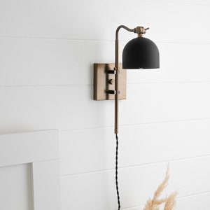 Op de afbeelding: Een zwarte en messing wandlamp met een zwarte kap. De lamp heeft een lang, zwart snoer.
