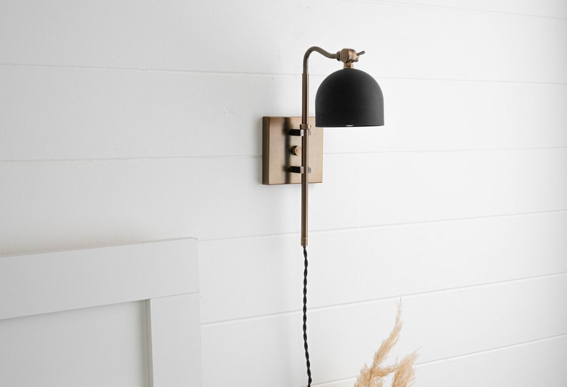 Op de afbeelding: Een zwarte en messing wandlamp met een zwarte kap. De lamp heeft een lang, zwart snoer.