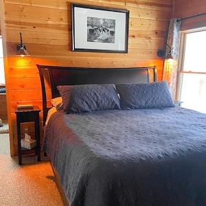 K&ouml;nnte beinhalten: Schlafzimmer mit dunkelgrauer Bettdecke und passenden Kissen. Das Bett hat ein schwarzes Kopfteil und steht vor einer holzget&auml;felten Wand. Ein gerahmtes Schwarz-Wei&szlig;-Foto h&auml;ngt &uuml;ber dem Bett. Zwei Wandleuchten flankieren das Bett, und ein kleiner Nachttisch ist sichtbar.