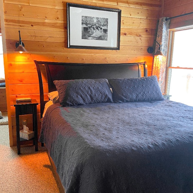 K&ouml;nnte beinhalten: Schlafzimmer mit dunkelgrauer Bettdecke und passenden Kissen. Das Bett hat ein schwarzes Kopfteil und steht vor einer holzget&auml;felten Wand. Ein gerahmtes Schwarz-Wei&szlig;-Foto h&auml;ngt &uuml;ber dem Bett. Zwei Wandleuchten flankieren das Bett, und ein kleiner Nachttisch ist sichtbar.