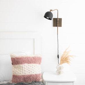 Op de afbeelding: Een witte slaapkamer met een bed met twee kussens, een roze en wit en een beige. Een wit nachtkastje met een vaas met gedroogd pampasgras en een zwarte en gouden wandlamp.