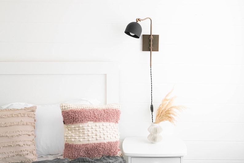 Op de afbeelding: Een witte slaapkamer met een bed met twee kussens, een roze en wit en een beige. Een wit nachtkastje met een vaas met gedroogd pampasgras en een zwarte en gouden wandlamp.