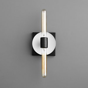 May include: A wall-mounted light fixture featuring a long, cylindrical, clear glass Edison-style bulb held by a central black socket. The fixture includes a circular white plate mounted onto a square black base, creating a layered geometric design.