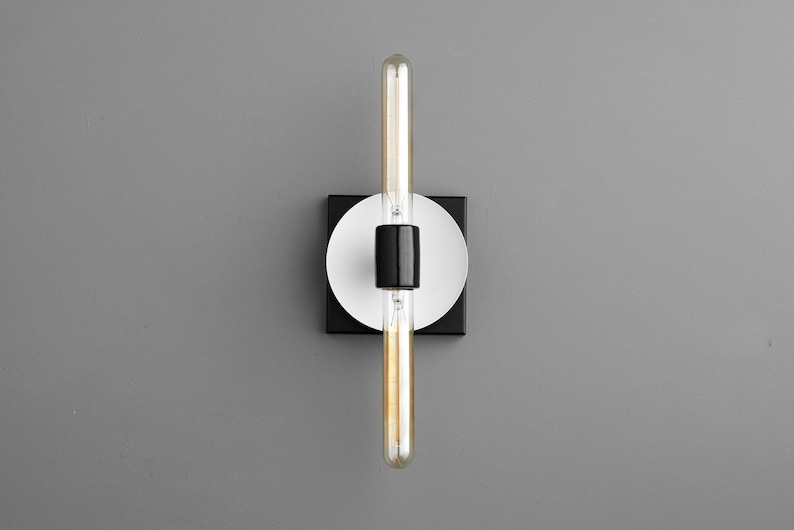 May include: A wall-mounted light fixture featuring a long, cylindrical, clear glass Edison-style bulb held by a central black socket. The fixture includes a circular white plate mounted onto a square black base, creating a layered geometric design.