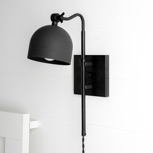Op de afbeelding: Zwarte wandlamp met een koepelvormige kap en een lange, verstelbare arm. De lamp is gemonteerd op een vierkante basis.