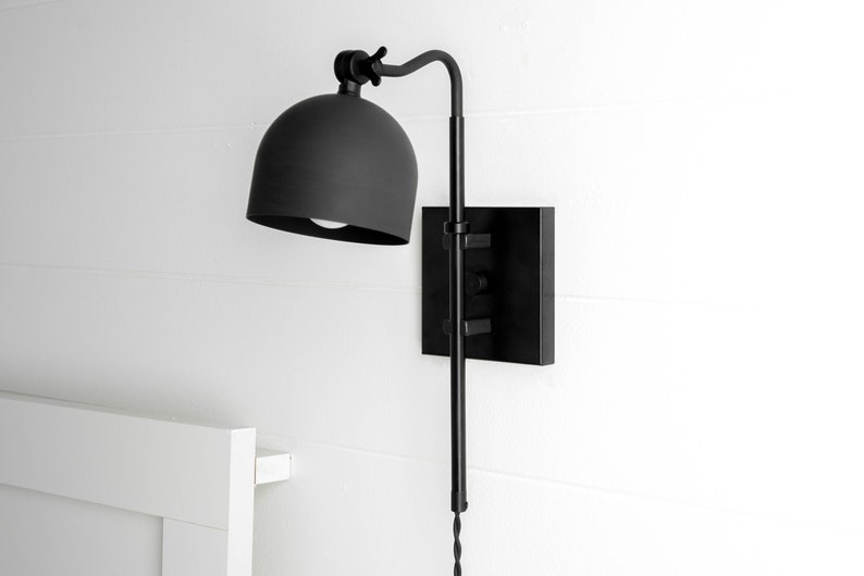 Op de afbeelding: Zwarte wandlamp met een koepelvormige kap en een lange, verstelbare arm. De lamp is gemonteerd op een vierkante basis.