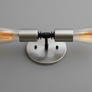 Op de afbeelding: Een wandlamp van geborsteld nikkel met twee zichtbare gloeilampen. De lamp heeft een zwarte metalen buis die de twee gloeilampen verbindt.