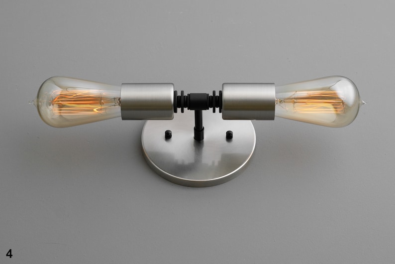 Op de afbeelding: Een wandlamp van geborsteld nikkel met twee zichtbare gloeilampen. De lamp heeft een zwarte metalen buis die de twee gloeilampen verbindt.