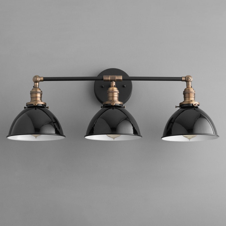 Puede incluir: Un aplique de ba&ntilde;o negro de tres luces con acabado en lat&oacute;n cepillado y pantallas de metal negro. El aplique tiene un dise&ntilde;o de barra recta con tres luces espaciadas uniformemente.