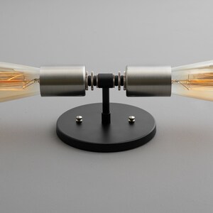 Op de afbeelding: Een zwarte en zilveren wandlamp met twee gloeilampen. De lamp heeft een ronde basis en twee armen die naar de zijkanten uitsteken. De gloeilampen zijn Edison-lampen met een warme gloed.