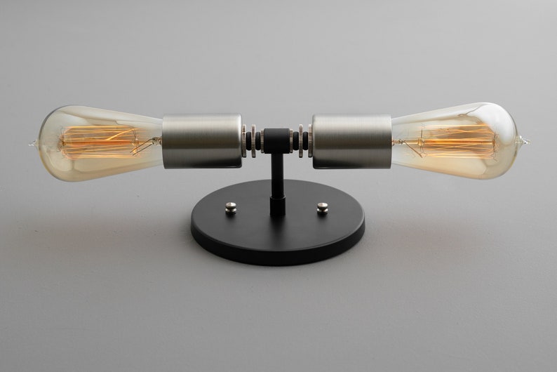 Op de afbeelding: Een zwarte en zilveren wandlamp met twee gloeilampen. De lamp heeft een ronde basis en twee armen die naar de zijkanten uitsteken. De gloeilampen zijn Edison-lampen met een warme gloed.