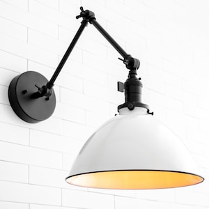 May include: A white and black industrial-style wall sconce with an adjustable arm. The sconce has a white metal shade with a gold interior.