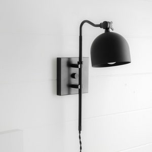 Op de afbeelding: Zwarte wandlamp met een koepelvormige kap en een lange, verstelbare arm. De lamp heeft een vierkante basis en een eenvoudig, modern ontwerp.