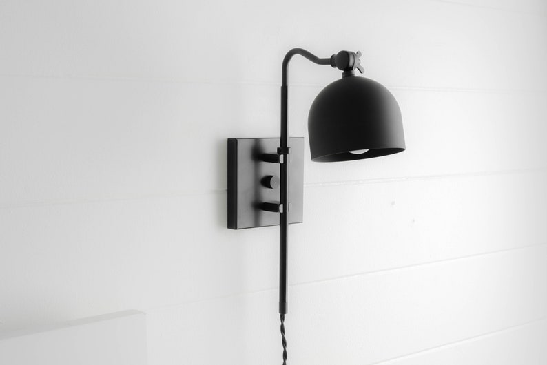 Op de afbeelding: Zwarte wandlamp met een koepelvormige kap en een lange, verstelbare arm. De lamp heeft een vierkante basis en een eenvoudig, modern ontwerp.