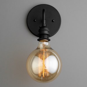 K&ouml;nnte beinhalten: Eine schwarze Wandleuchte mit einer runden R&uuml;ckplatte und einer einzelnen Gl&uuml;hbirne. Die Gl&uuml;hbirne ist eine klare Glaskugel mit einem sichtbaren Filament.