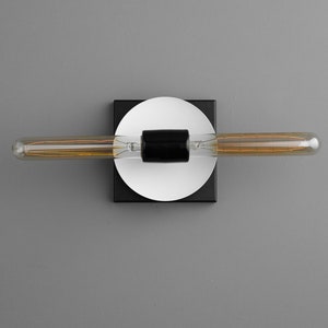 May include: A wall-mounted light fixture featuring a long, horizontal tubular Edison bulb with visible internal filaments. The bulb is centered on a black cylindrical socket, which is mounted onto a white circular plate and a black square base.