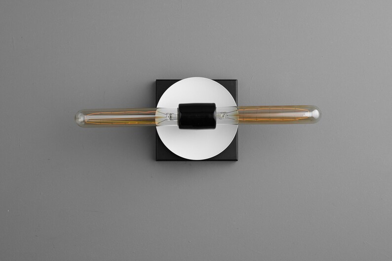 May include: A wall-mounted light fixture featuring a long, horizontal tubular Edison bulb with visible internal filaments. The bulb is centered on a black cylindrical socket, which is mounted onto a white circular plate and a black square base.