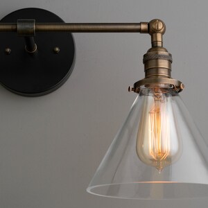 May include: A vintage-style wall sconce with a clear glass shade and a warm, glowing Edison bulb. The fixture has a bronze finish and a black mounting plate. The design is industrial and suitable for home lighting.