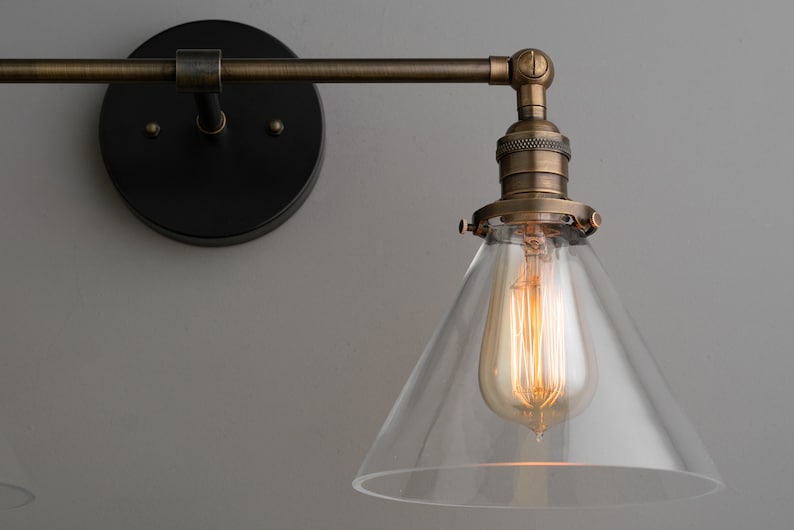 May include: A vintage-style wall sconce with a clear glass shade and a warm, glowing Edison bulb. The fixture has a bronze finish and a black mounting plate. The design is industrial and suitable for home lighting.