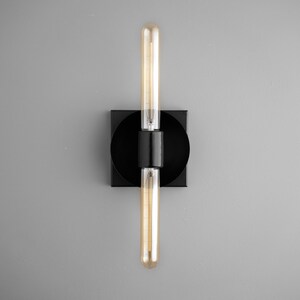 May include: A wall sconce featuring a matte black square base with a circular center mount. Two long, cylindrical, amber-tinted Edison-style light bulbs are positioned vertically, one extending upward and one downward from the central black fixture.