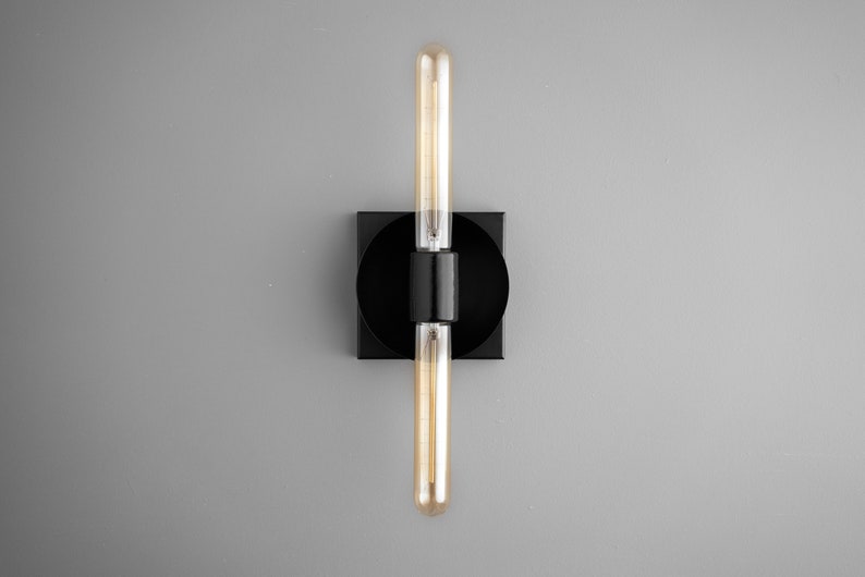 May include: A wall sconce featuring a matte black square base with a circular center mount. Two long, cylindrical, amber-tinted Edison-style light bulbs are positioned vertically, one extending upward and one downward from the central black fixture.