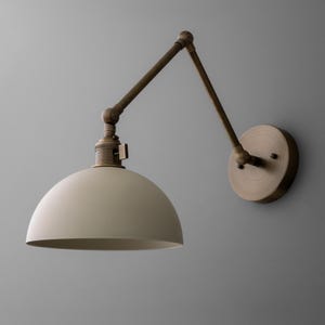 May include: A wall-mounted, adjustable lamp with a cream-colored, dome-shaped shade. The lamp features a bronze-colored, articulated arm and a circular wall plate. The design is reminiscent of industrial or vintage lighting.