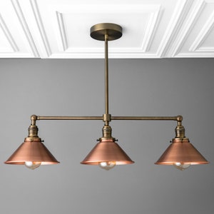 May include: A three-light pendant light fixture with a bronze finish. The fixture has three copper-colored cone-shaped shades, each housing a lightbulb. The fixture is suspended from the ceiling by a bronze rod.