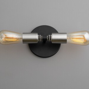 Op de afbeelding: Een zwarte en zilveren wandlamp met twee Edison-lampen. De lamp heeft een ronde zwarte basis en twee zilveren armen die naar de zijkanten uitsteken. De lampen zijn van helder glas met een warm wit licht.