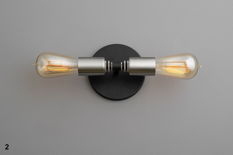 Op de afbeelding: Een zwarte en zilveren wandlamp met twee Edison-lampen. De lamp heeft een ronde zwarte basis en twee zilveren armen die naar de zijkanten uitsteken. De lampen zijn van helder glas met een warm wit licht.