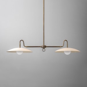 May include: A brass two-light pendant light fixture with white shades. The fixture has a long, straight rod with two arms extending out to the sides, each holding a white shade. The shades are round and slightly curved, and they have a white, opaque finish.