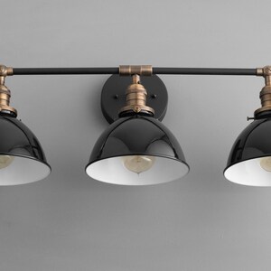 Puede incluir: Una l&aacute;mpara de pared de tres luces negra y de lat&oacute;n con pantallas de metal negro. Las pantallas tienen un interior blanco y est&aacute;n inclinadas hacia abajo.
