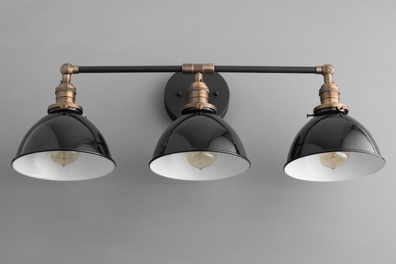 Puede incluir: Una l&aacute;mpara de pared de tres luces negra y de lat&oacute;n con pantallas de metal negro. Las pantallas tienen un interior blanco y est&aacute;n inclinadas hacia abajo.