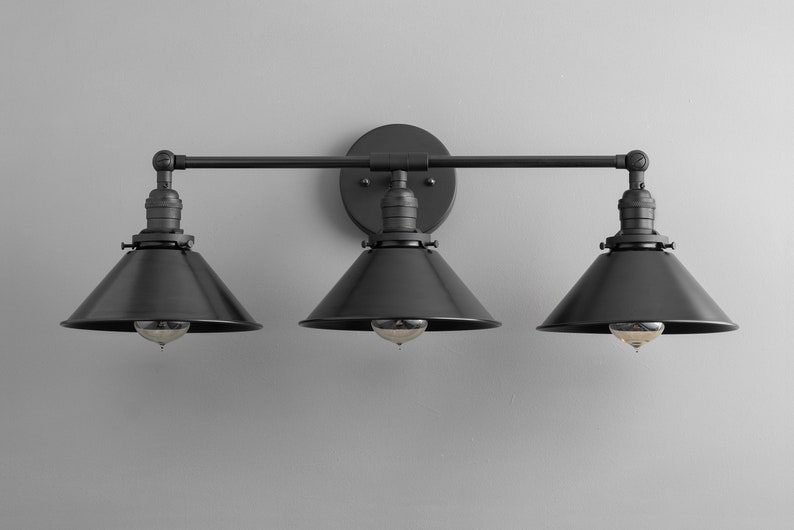 May include: Black three-light vanity light fixture with black metal shades. The fixture has a straight bar design with three arms extending out from a central wall mount.