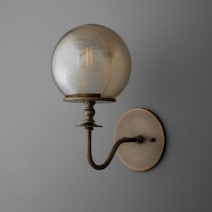 May include: A wall-mounted light fixture with a spherical, amber-colored glass shade. The fixture has a bronze finish, including a round backplate, a curved arm, and a decorative base supporting the globe. The light bulb is visible inside the glass.