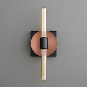 May include: A wall sconce featuring a long, tubular Edison-style light bulb mounted vertically. The fixture includes a central black cylindrical socket, a circular copper-toned backplate, and a square black base plate, creating a layered design.