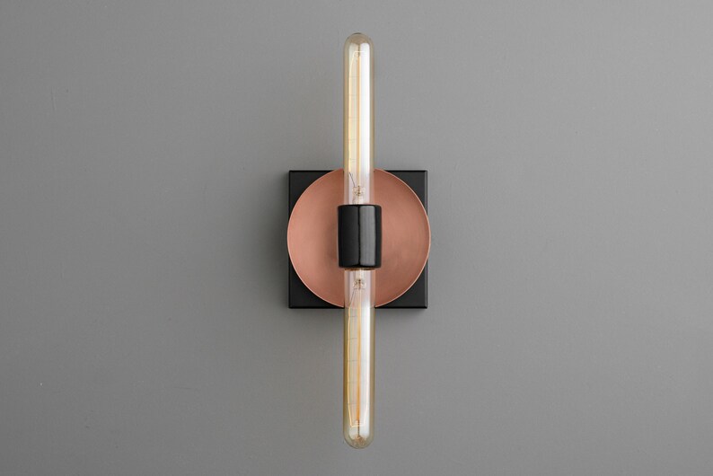 May include: A wall sconce featuring a long, tubular Edison-style light bulb mounted vertically. The fixture includes a central black cylindrical socket, a circular copper-toned backplate, and a square black base plate, creating a layered design.