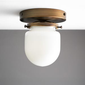 May include: A ceiling-mounted light fixture with a brass-colored base and a white, rounded glass shade. The fixture is designed to be mounted flush against the ceiling, providing a clean and modern look. The glass shade diffuses light evenly.