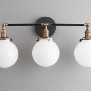 May include: A three-light vanity fixture with a black metal frame and white glass globes. The fixture has a brushed brass finish.
