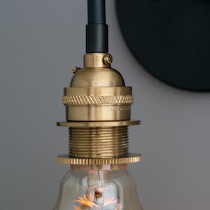 K&ouml;nnte beinhalten: Nahaufnahme einer Messing-Leuchte mit schwarzem Kabel und einer klaren Gl&uuml;hbirne. Die Leuchte hat ein dekoratives Gewinde-Design und ist an einer schwarzen Wandplatte befestigt.