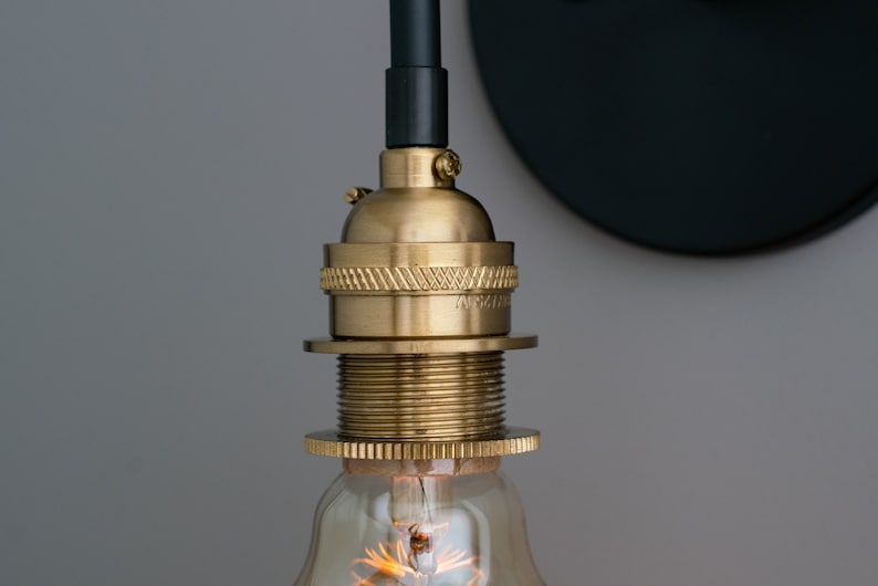K&ouml;nnte beinhalten: Nahaufnahme einer Messing-Leuchte mit schwarzem Kabel und einer klaren Gl&uuml;hbirne. Die Leuchte hat ein dekoratives Gewinde-Design und ist an einer schwarzen Wandplatte befestigt.